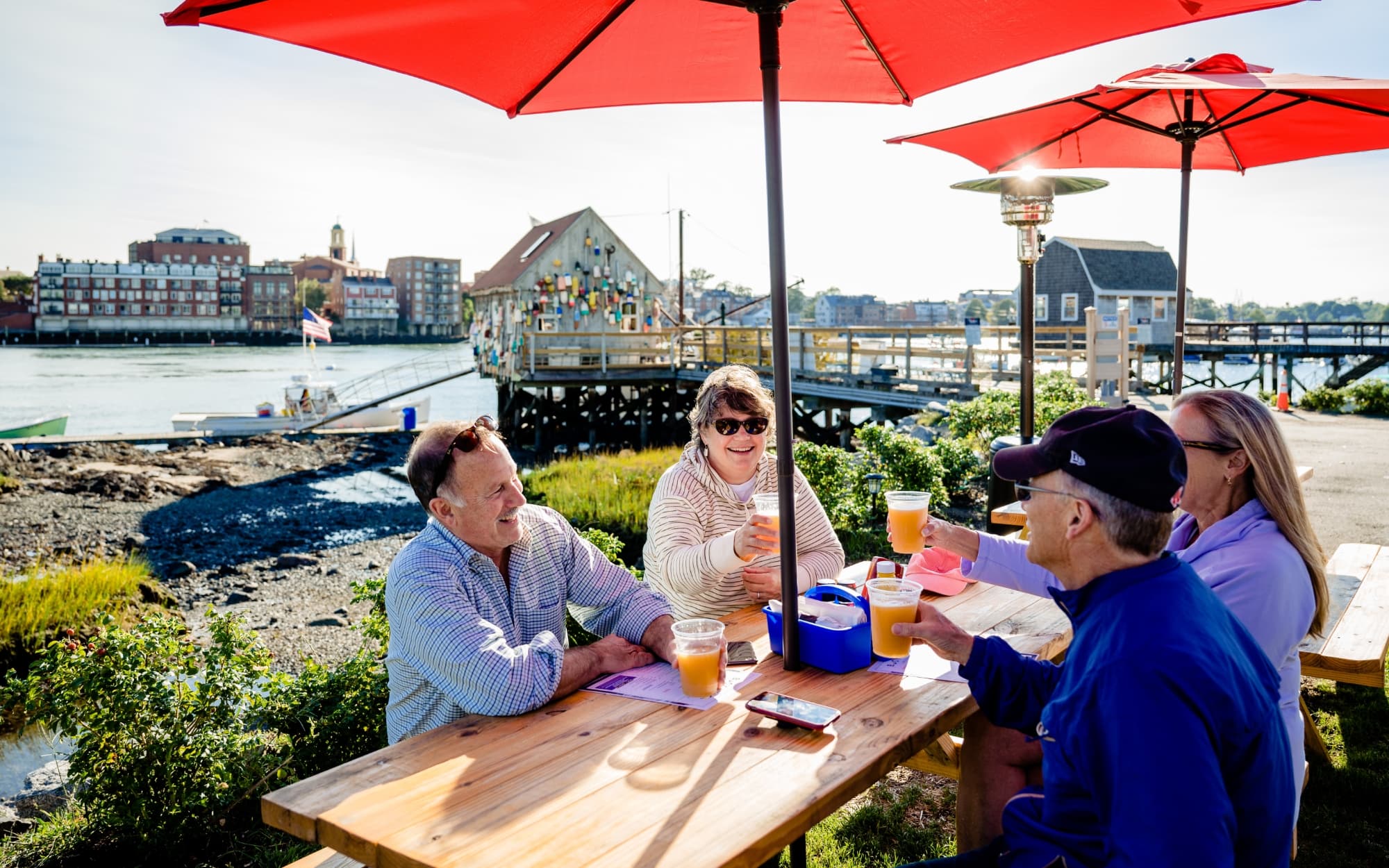 Four patrons are smiling while seated outside in front of an ocean view at the Buoy Shack. They are offering cheers with their beverages.